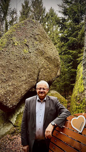 Ein Mann steht lächelnd vor einer großen, moosbedeckten Felsformation in einem Wald. Er lehnt an einer Holzbank mit einem herzförmigen Schild, das einen freundlichen Akzent in der natürlichen Umgebung setzt.