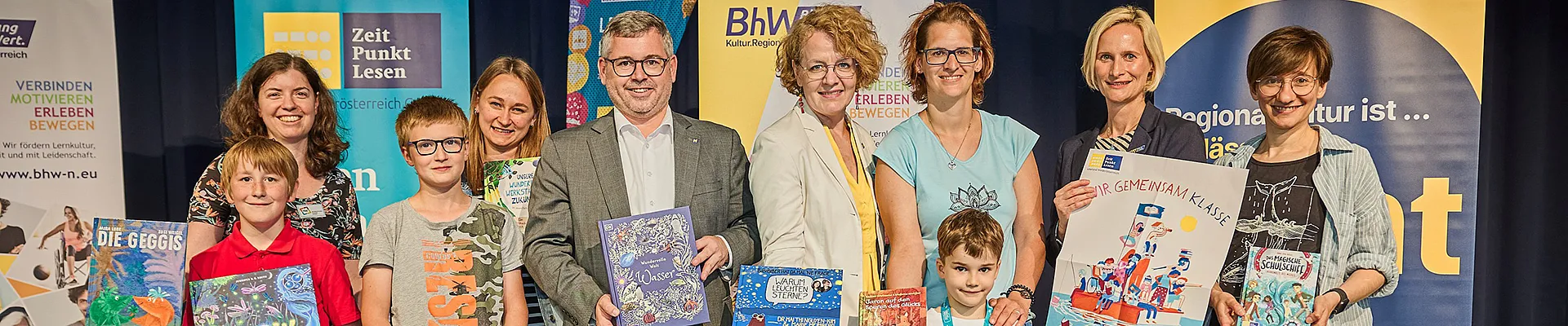 Gruppenbild, in der ersten Reihe stehen Kinder und in der zweiten Reihe Erwachsene Personen. Im Hintergrund stehen einige Rollups vom BhW, Zeit Punkt Lesen und Kulturregion.
