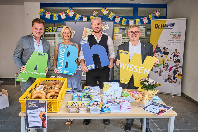 Gruppenbild der vier Personen. Sie halten große Buchstaben in der Hand. davor steht ein Tisch mit Infomaterialien vom BhW.