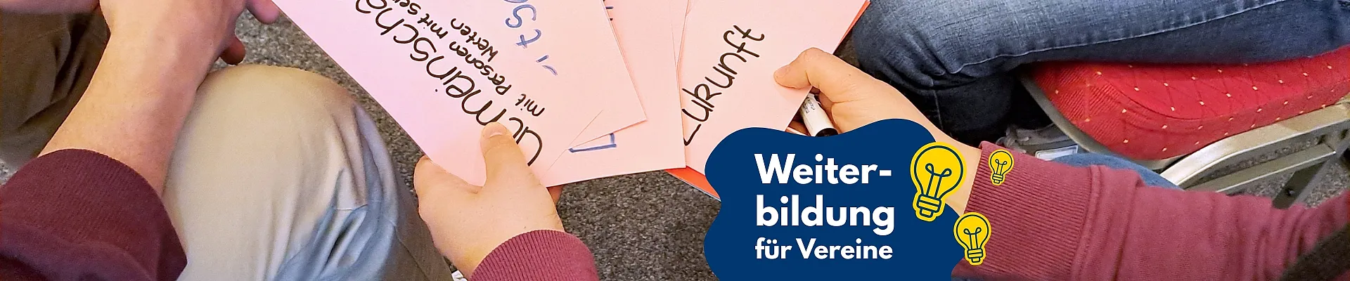 Auf dem Bild sitzen mehrere Personen zusammen und halten beschriftete Karten in den Händen, auf denen Begriffe wie „Zukunft“ und „Umgestaltung“ stehen. Rechts unten ist ein grafisches Element mit der Aufschrift „Weiterbildung für Vereine“ und kleinen Glühbirnen-Symbolen zu sehen.