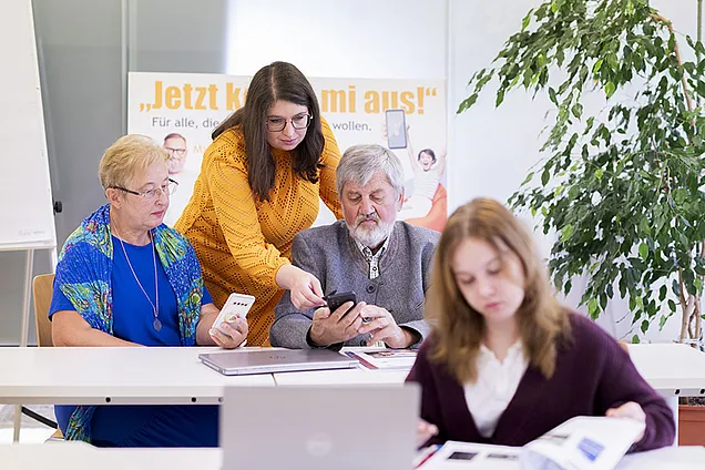 Eine junge Frau unterstützt zwei ältere Personen beim Umgang mit dem Smartphone, während eine Schülerin im Vordergrund konzentriert in den Lehrunterlagen blättert. Die Szene spielt sich in einem modernen Lernumfeld ab, das digitale Bildung für alle Generationen fördert.