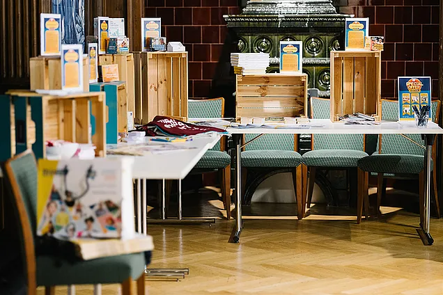 Folder und Werbemittel stehen in Holzboxen auf Tischen. Sie erinnern an einen Supermarkt.