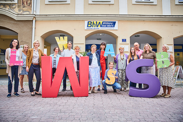 Die Lernbegleiterinnen und Lernbegleiter der Basisbildung stehen vor dem Haus Linzerstraße 7. Davor stehen noch zwei sehr große Buchstaben aus Schaumstoff. Ein großes "W" in rot und ein großes "S" in lila.  Einige Personen halten auch kleinere Buchstaben in den Händen.