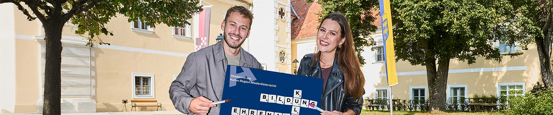 Zwei junge Erwachsene sitzen im Freien vor einem historischen Gebäude und halten gemeinsam ein Schild mit einem Kreuzworträtsel-Design. Darauf stehen die Begriffe „Bildung“, „Kultur“ und „Ehrenamt“ sowie der Hinweis auf die Akademie der Kultur.Region.Niederösterreich.