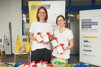 BhW Geschäftsführerin Melanie Winkler und Basisbildungs-Mitarbeiterin Claudia Geist. Sie halten kleine Sackerl mit Buchstaben-Nudeln und einem Info-Zettel über den Weltalphabetisierungstag in den Händen.