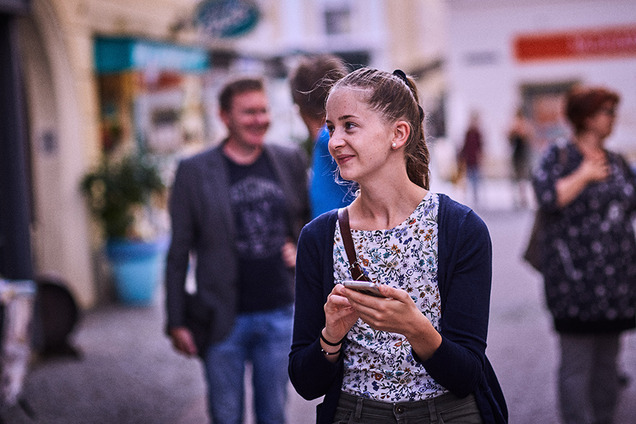 Eine junge Frau geht durch die Straßen von St. Pölten, hält ihr Smartphone in der Hand und schaut sich neugierig um.