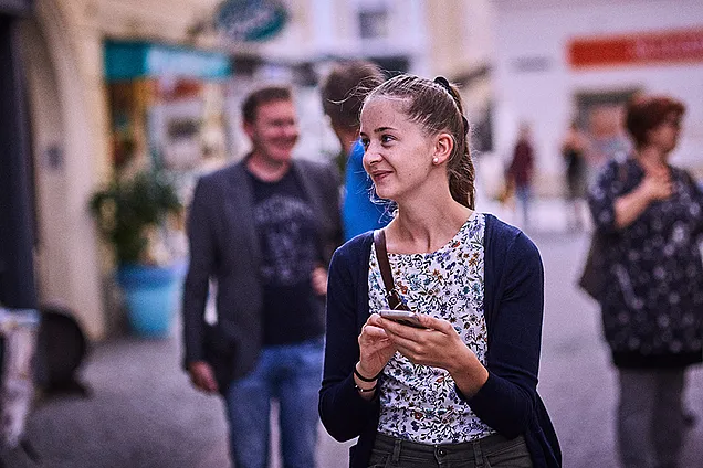Eine junge Frau geht durch die Straßen von St. Pölten, hält ihr Smartphone in der Hand und schaut sich neugierig um.