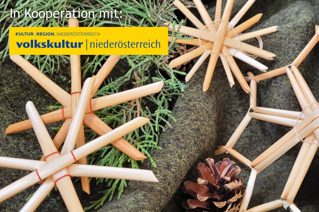 Strohsterne liegend auf einem grauen Hintergrund. Daneben liegen ein paar grüne Zweige und ein kleiner Tannenzapfen.