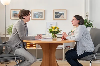 Zwei Frauen sitzen sich an einem runden Tisch in einem hellen Büro gegenüber und führen ein Beratungsgespräch. Im Hintergrund sind Bilder an der Wand, ein Blumenstrauß auf dem Tisch und Arbeitsmaterialien zu sehen, was eine ruhige, professionelle Atmosphäre vermittelt.