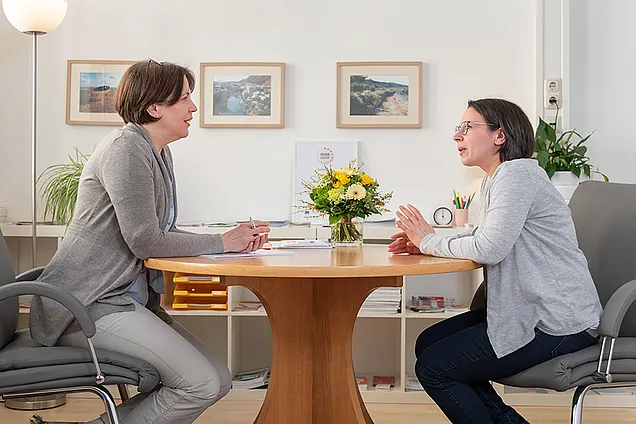Zwei Frauen sitzen sich an einem runden Tisch in einem hellen Büro gegenüber und führen ein Beratungsgespräch. Im Hintergrund sind Bilder an der Wand, ein Blumenstrauß auf dem Tisch und Arbeitsmaterialien zu sehen, was eine ruhige, professionelle Atmosphäre vermittelt.