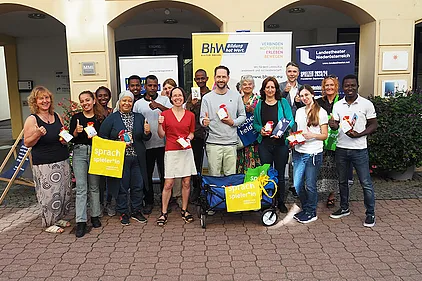 Gruppenbild auf der Straße vor dem BhW. Man sieht eine Gruppe von 16 Personen. Dabei sind Trainer/innen der Basisbildung, Schauspieler Sven Kaschte und Passanten. Sie halten kleine Sackerl mit Buchstaben-Nudeln und einem Info-Zettel über den Weltalphabetisierungstag in den Händen.