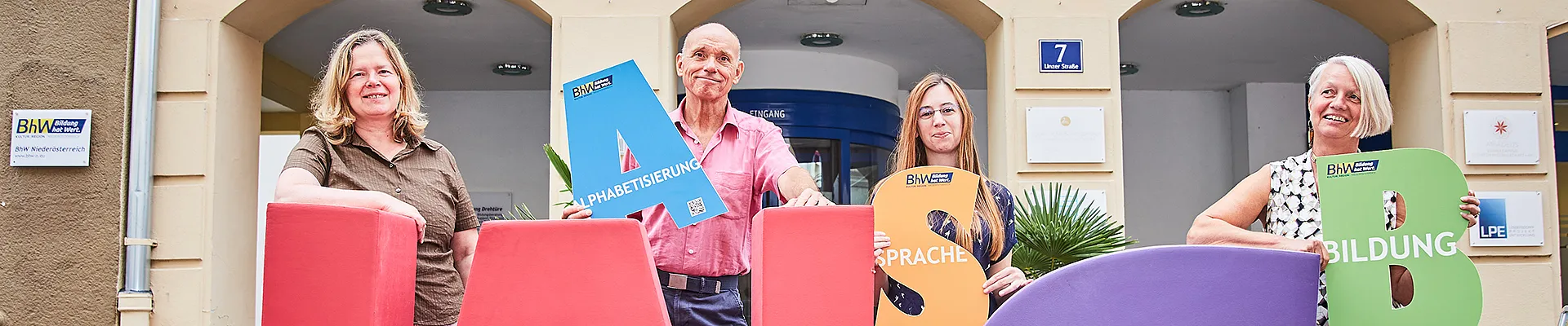 Vier Lernbegleiterinnen und Lernbegleiter der Basisbildung stehen vor dem Haus Linzerstraße 7. Davor stehen noch zwei sehr große Buchstaben aus Schaumstoff. Ein großes "W" in rot und ein großes "S" in lila. Drei der Personen halten auch kleinere Buchstaben in den Händen.