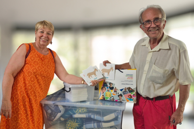 Eine Frau mit Sommerkleid und ein Herr mit kurzer Hose und Hemd stehen vor einer Box mit Materialien. Sie halten ein Spiel