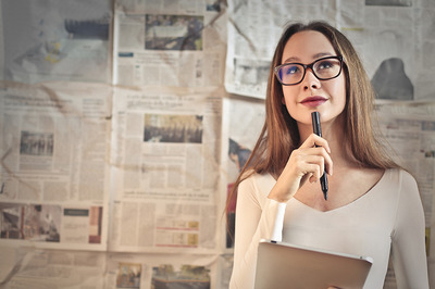 Frau mit langen hellbraunen Haaren und einer Brille, die nachdenklich schaut und sich einen Kugelschreiber an den Mund hält. Im Hintergrund sind Zeitungsseiten zu sehen.