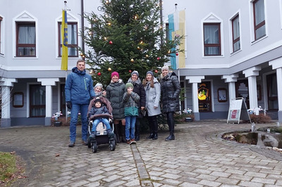 Das Bild zeigt den Innenhof des Gemeindeamts in Kirchberg am Wechsel. Eine Familie mit Freunden steht vor einem festlich geschmückten Christbaum. Alle Personen lächeln und freuen sich auf Weihnachten.