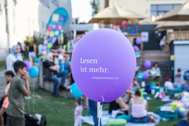Lesepicknick im Freien. Kinder und Erwachsene sitzen auf Picknickdecken. Auf einem lilafarbenen Luftballon steht „Lesen ist mehr“.