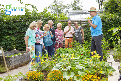 Das Bild zeigt eine Personengruppe, die bei einem Pflanzenbeet steht und von einem Gärtner essbare Blüten zum Verkosten bekommt.