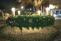 Das Foto zeigt einen großen Adventkranz aus Tannenzweigen mit bunten Weihnachtskugeln und vier leuchtenden Kerzen darauf. Die Fotoaufnahme entstand abends.