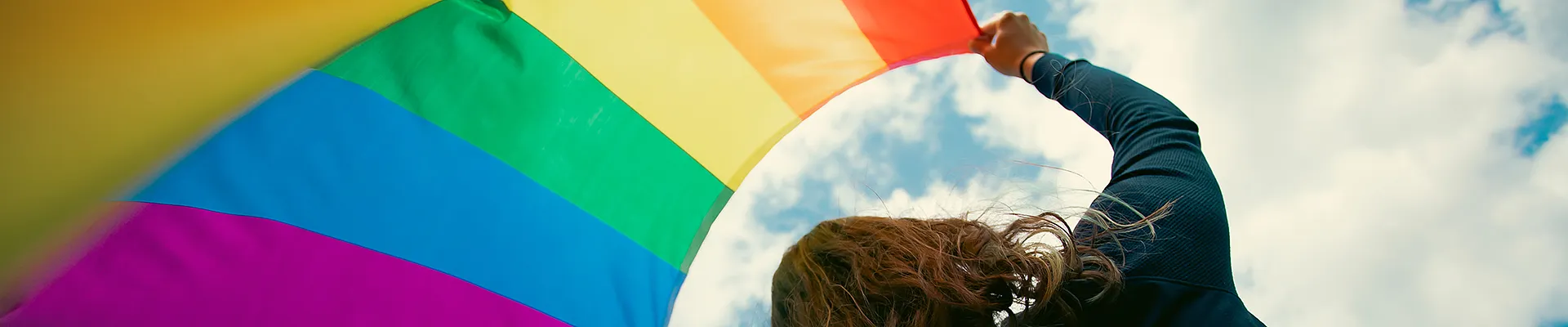 Eine Foto einer Frau die eine Regenbogenfahne in den Himmel hält.