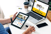 Männerhände halten ein Tablet auf dem die Startseite von https://www.bildungsangebote.at zu sehen ist. Dahinter steht ein Laptop auf einem Schreibtisch, auf diesem sieht man die Startseite der Homepage https://www.bildungsgemeinden-noe.at