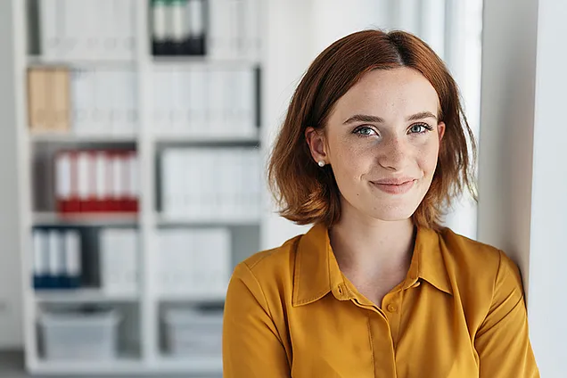 Portraitfoto einer jungen Frau mit roten Haaren. Sie trägt eine dottergelbe Bluse und lächelt in die Kamera. Im Hintergrund sieh tman ein Bücherregal.