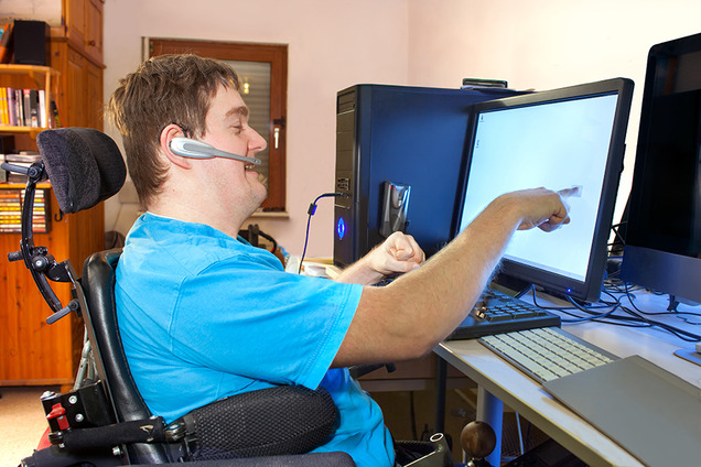 Ein blinder junger Mann, der im Rollstuhl sitzt, bedient seinen Computer mit Hilfe eines TouchScreens.