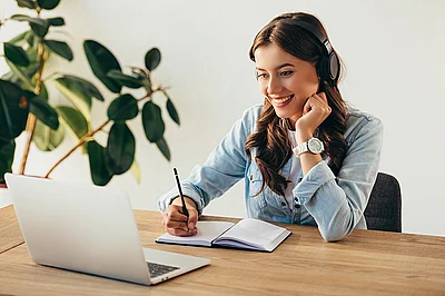 Eine Frau mit Kopfhörern vor einem Laptop macht sich Notizen