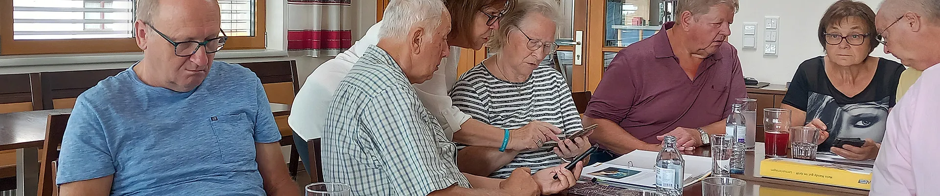Mehrere Personen sitzen an einem Tisch und sind in ihr Smartphone vertieft, eine Person erklärt etwas.