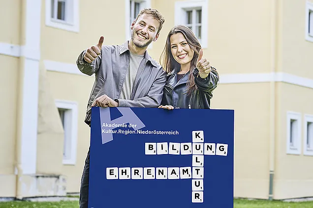 Ein junger Mann und eine junge Frau stehen lächelnd nebeneinander vor einem Gebäude und zeigen beide mit dem Daumen nach oben. Vor ihnen halten sie ein großes Schild mit einem Kreuzworträtsel-Design, auf dem die Begriffe „Bildung“, „Kultur“ und „Ehrenamt“ sowie der Schriftzug „Akademie der Kultur.Region.Niederösterreich“ zu sehen sind.