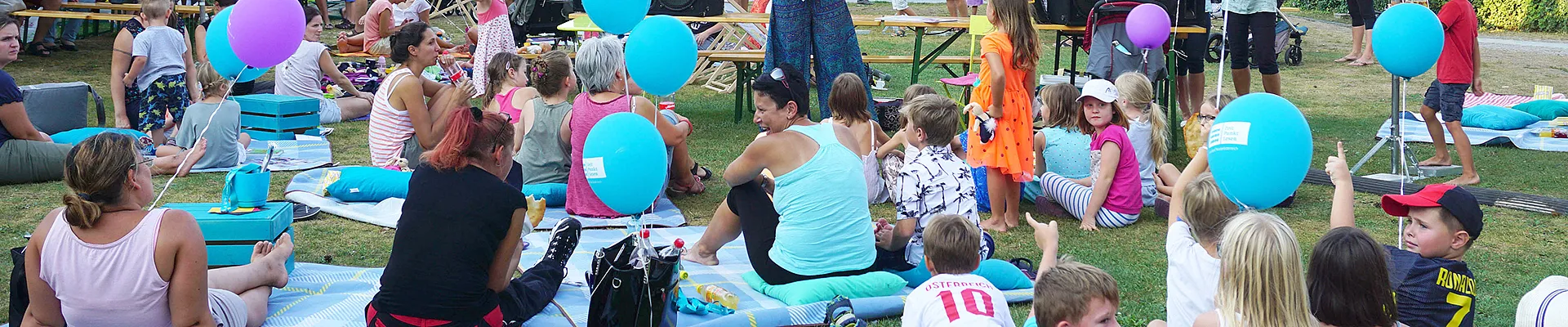 Auf dem Bild ist das Setting eines Lesepicknicks im Freien zu sehen, mit Picknickdecken, Kindern und ihren Eltern sowie eine Bühne für eine Lesung.