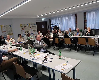 Foto von 3 langen Tischen welche in einer U Form aufgestellt sind. Alle Teilnehmer der Hauptregionskonferenz sitzen an diesen Tischen und schauen nach vorne in die Mitte. Ebenso befinden sich Zettel und Getränke auf den Tischen.