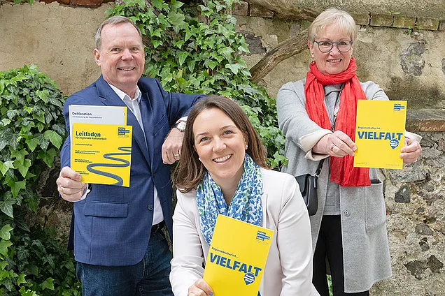 Drei Personen, ein Mann, zwei Frauen, halten jeweils eine gelbe Broschüre in der Hand. 