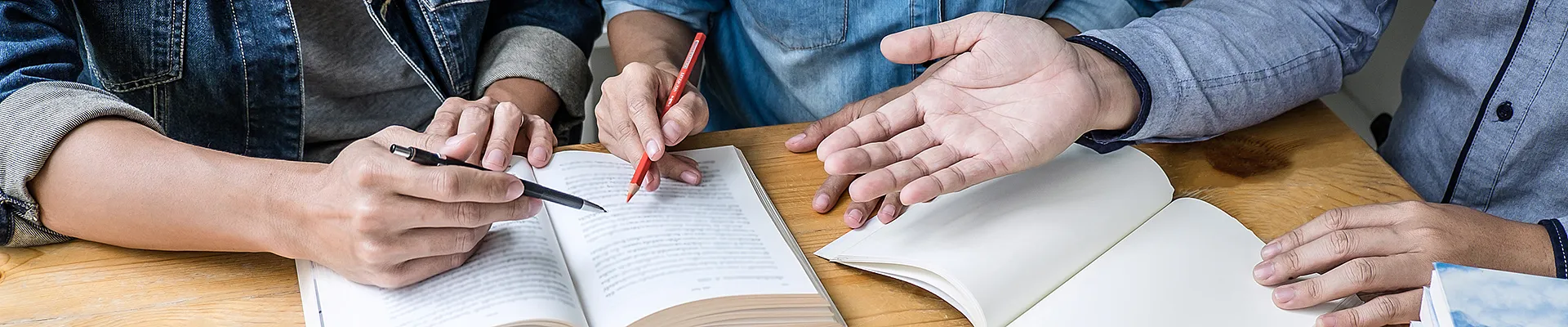 Auf einem Schreibtisch liegen aufgeschlagene Bücher und Stifte. Man sieht noch die Oberkörper und Amre von drei Personen die sich anscheinend angeregt unterhalten.