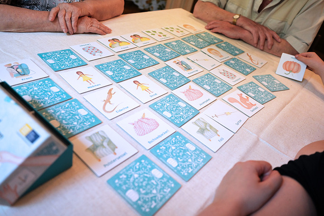 Die Karten des Mundart-Memospieles sind auf einem Tisch aufgebreitet. Die meisten Karten sind verdeckt. Nur ein paar Karten liegen offen auf dme Tisch. Rund um den Tisch sitzt eine Familie beim Spiele-Nachmittag.
