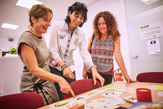Drei fröhliche Lernbegleiterinnen der Basisbildung. Sie stehen vor einem Tisch auf dem Kärtchen mit Tiermotiven liegen.