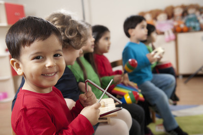Ein paar Kinder probieren Instrumente aus, im Vordergrund auf der linken Seite hält ein Junge im roten Pullover eine Triangel und lächelt in die Kamera.