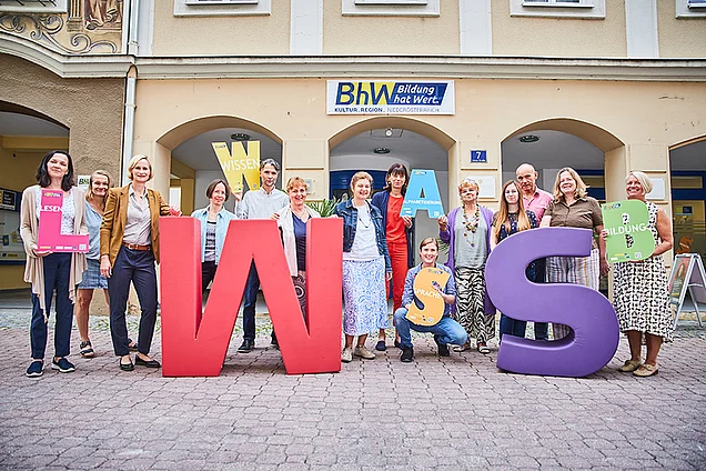 Die Lernbegleiterinnen und Lernbegleiter der Basisbildung stehen vor dem Haus Linzerstraße 7. Davor stehen noch zwei sehr große Buchstaben aus Schaumstoff. Ein großes "W" in rot und ein großes "S" in lila.  Einige Personen halten auch kleinere Buchstaben in den Händen.