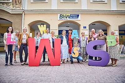 Die Lernbegleiterinnen und Lernbegleiter der Basisbildung stehen vor dem Haus Linzerstraße 7. Davor stehen noch zwei sehr große Buchstaben aus Schaumstoff. Ein großes "W" in rot und ein großes "S" in lila. Einige Personen halten auch kleinere Buchstaben in den Händen.
