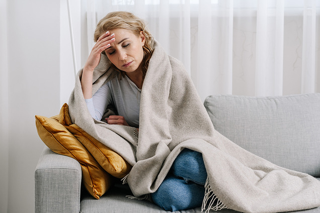 Eine Frau sitzt in eine Decke gehüllt auf einem Sofa und greift sich erschöpft an die Stirn.