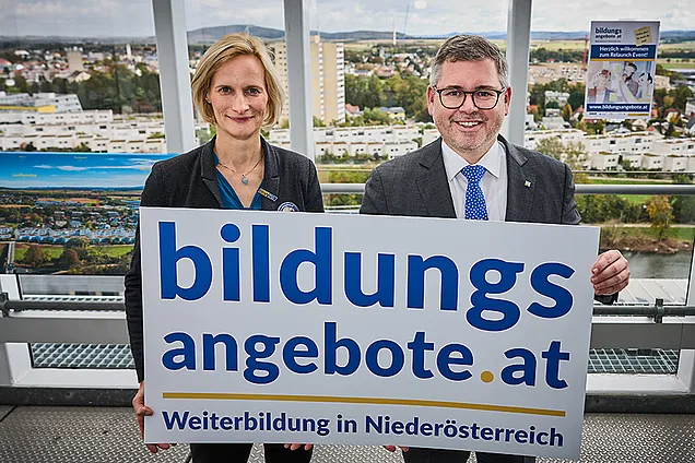 Therese Reinel und Landesrat Ludwig Schleritzko halten auf der Aussichtsterrasse vom Klangturm St. Pölten ein Schild mit der Aufschrift bildungsangebote.at hoch.