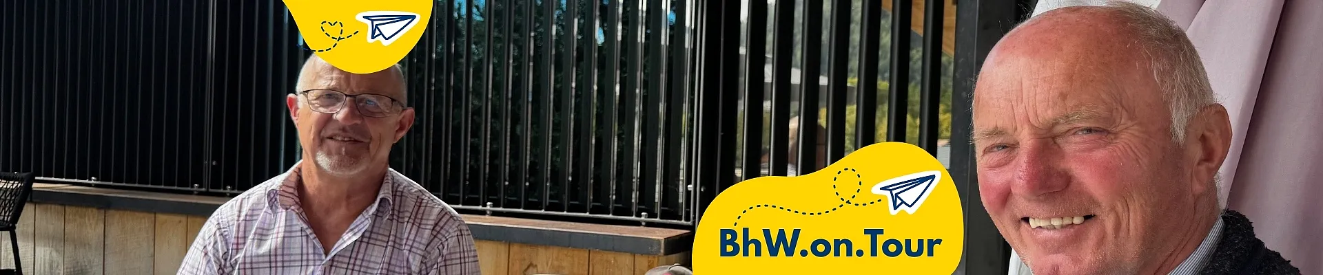 Zwei Männer sitzen lächelnd an einem Tisch in einem sonnigen Außenbereich eines Cafés und haben BhW-Unterlagen vor sich liegen. Im Vordergrund ist ein gelbes BhW.on.Tour-Grafikelement mit Papierflieger-Symbol eingeblendet.
