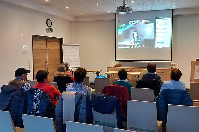 Mehrere Personen sitzen in einem Seminarraum und verfolgen eine Online-Präsentation, die auf eine Leinwand projiziert wird. Auf dem Bildschirm ist eine Frau im Videocall mit dem Logo von „BhW Niederösterreich“ zu sehen.