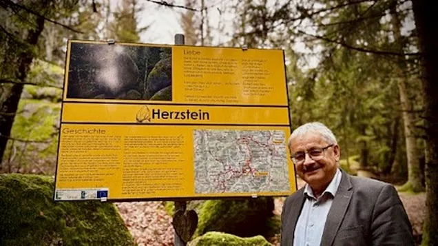 Ein Mann steht lächelnd neben einer großen gelben Informationstafel mit der Überschrift „Herzstein“, die Geschichte, Texte und eine Karte zeigt. Die Szene spielt in einem bewaldeten Gebiet, umgeben von moosigen Felsen und Bäumen.