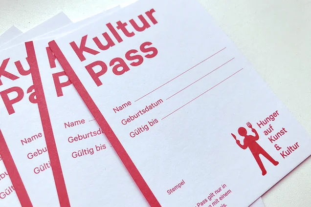Foto von mehreren Kulturpässen. Es steht auf einem ca. A7 großen Karton, in roter Schrift, Kulturpass, dann Felder zum Ausfüllen für Name und Geburtsdatum. Und man sieht das Logo: das besteht aus einem roten Männchen mit Messer und Gabel in der Hand und daneben steht: Hunger auf Kunst & Kultur