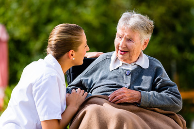 Eine ältere Frau in einem Rollstuhl sitzend und eine Pflegerin links neben ihr, die sich fürsorglich um sie kümmert.