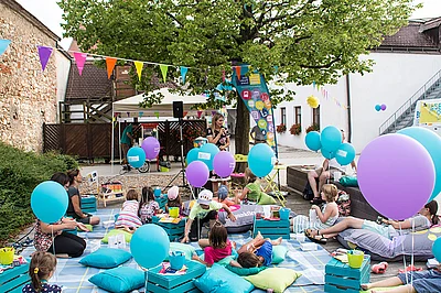 Kinder sitzen auf Decken und Pölter im Park und lauschen einer Geschichte.