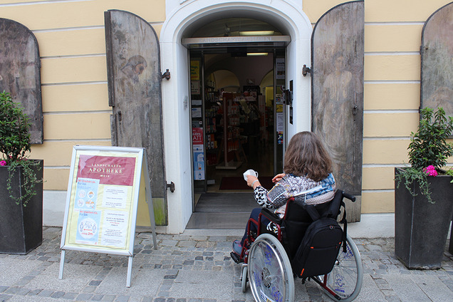 Eine Frau im Rollstuhl vor dem stufenlosen Eingang zu einer Apotheke