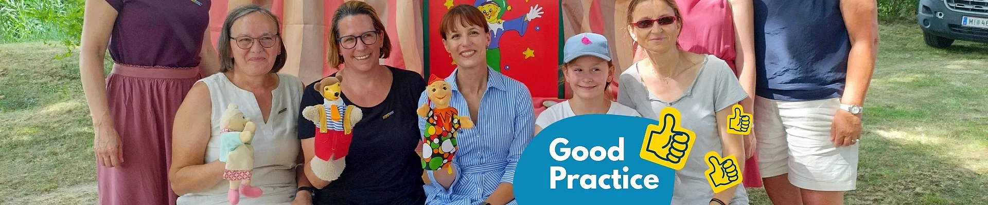 Eine Gruppe von acht Personen sitzt und steht vor einem bunten Zelt, das mit BhW-Wimpeln geschmückt ist, und einige halten Handpuppen in den Händen. Im linken unteren Bildbereich befindet sich ein blaues „Good Practice“-Grafikelement mit einem Daumen-hoch-Symbol.