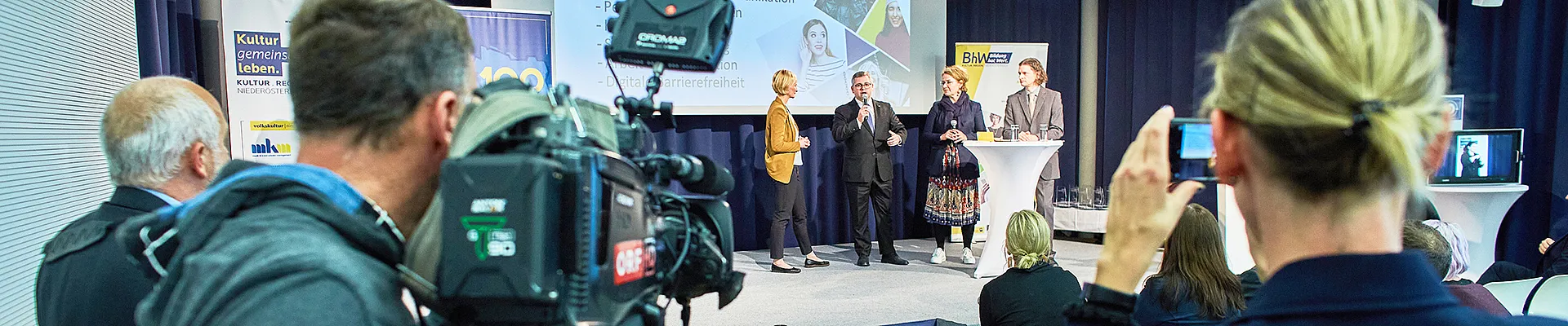 Im Vordergrund sieht man einen Mann mit einer großen Fernsehkamera und daneben eine Frau mit einem Handy mit dem sie filmt. beide filmen eine Interviewszene auf einer Bühne, auf der vier Personen stehen.