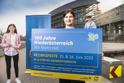 Eine Frau steht im Vordergrund mit einem Schild in der Hand, auf dem für die Bezirksfeste am 25. und 26. Juni geworben wird. Links davon steht eine junge Frau mit Querflöte und weiter im Hintergrund steht ein junger Mann mit Klarinettte. Das Foto wurde vor dem Landhaus St. Pölten aufgenommen.